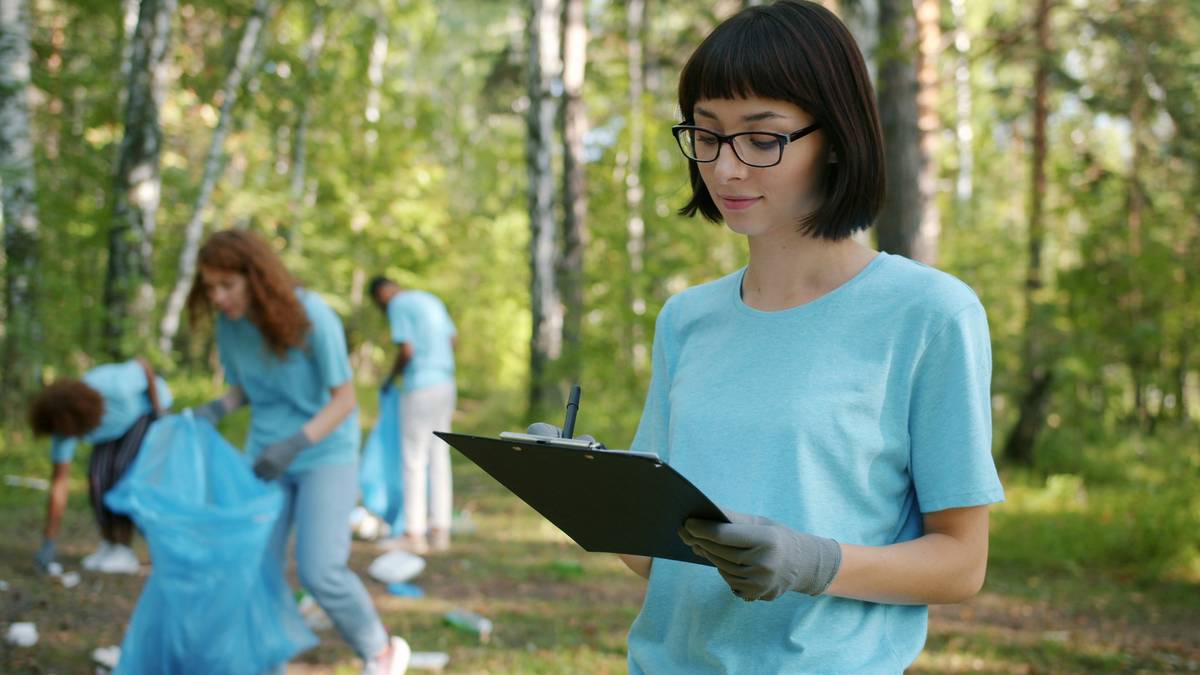 Portrait of young eco-friendly lady doing paperwork during forest clean-up holding folder while multi-ethnic team of activists in uniform is picking garbage.